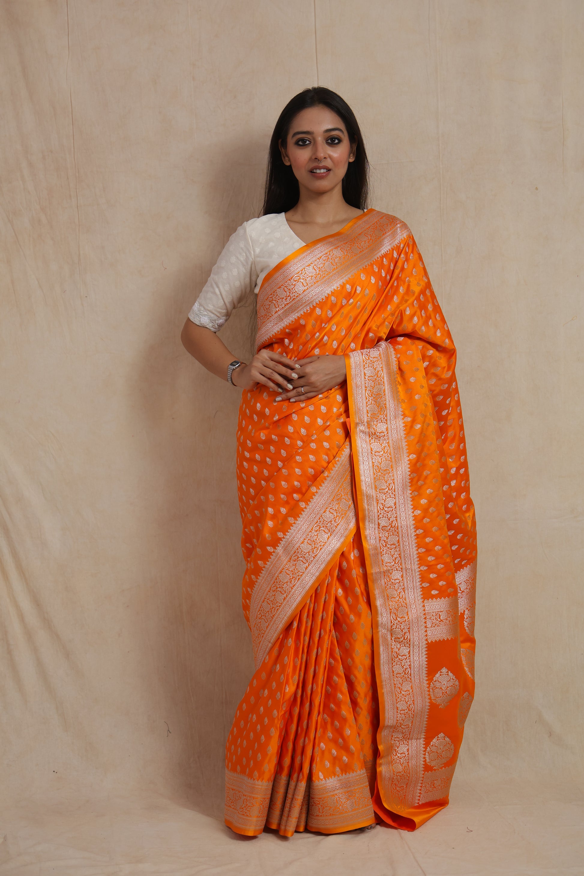 Model wearing vibrant orange Banarasi silk saree with silver zari buttas and woven border.