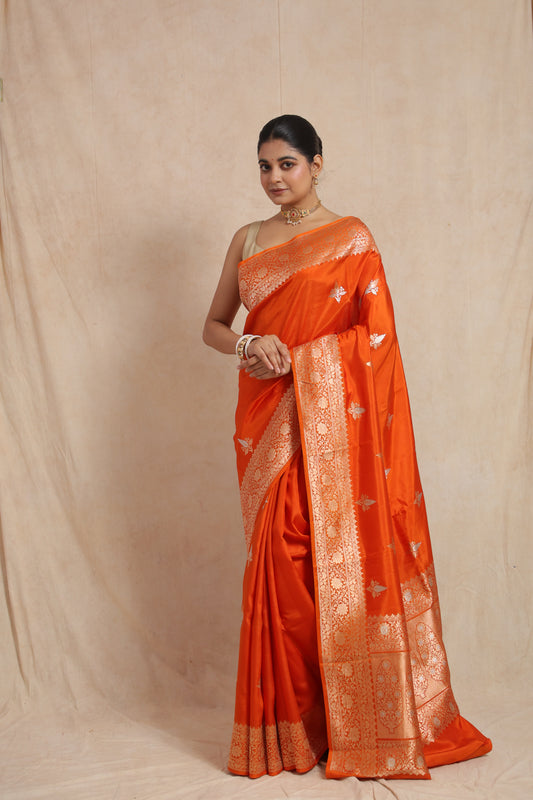 Front view of radiant tangerine orange Banarasi silk saree with floral woven border and traditional buttas